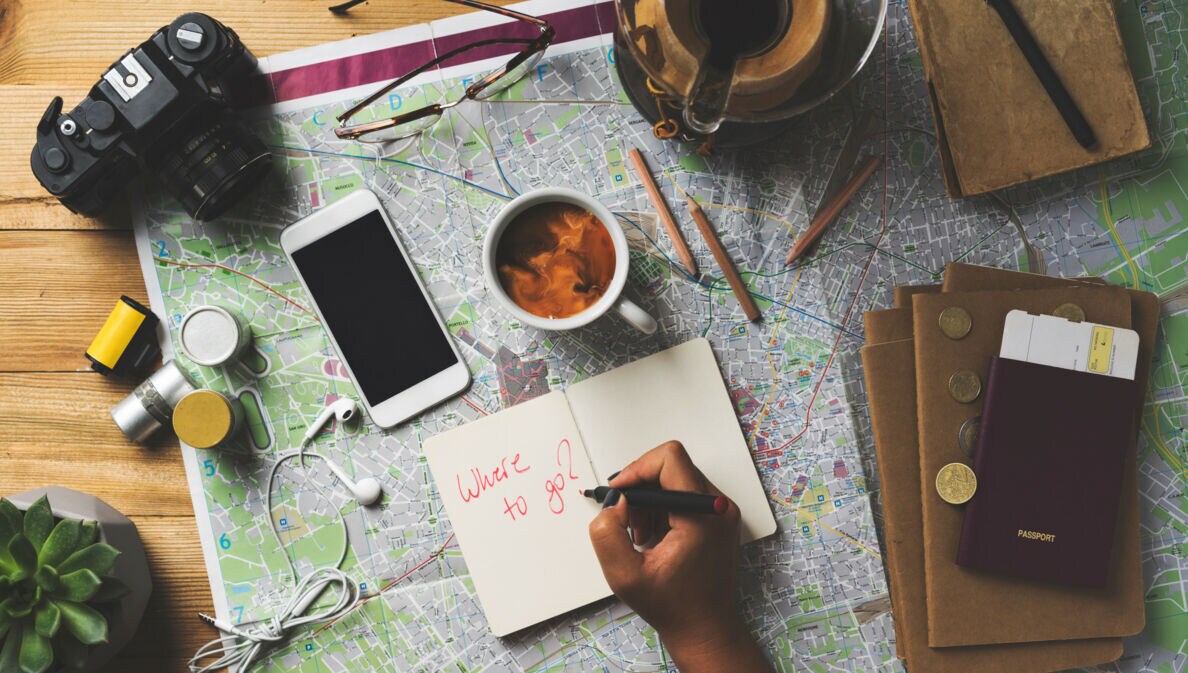 Hand schreibt 'Where to go?' in Notizbuch auf Landkarte mit Kamera, Smartphone, Kaffeetasse, Brille, Reisepass und Münzen