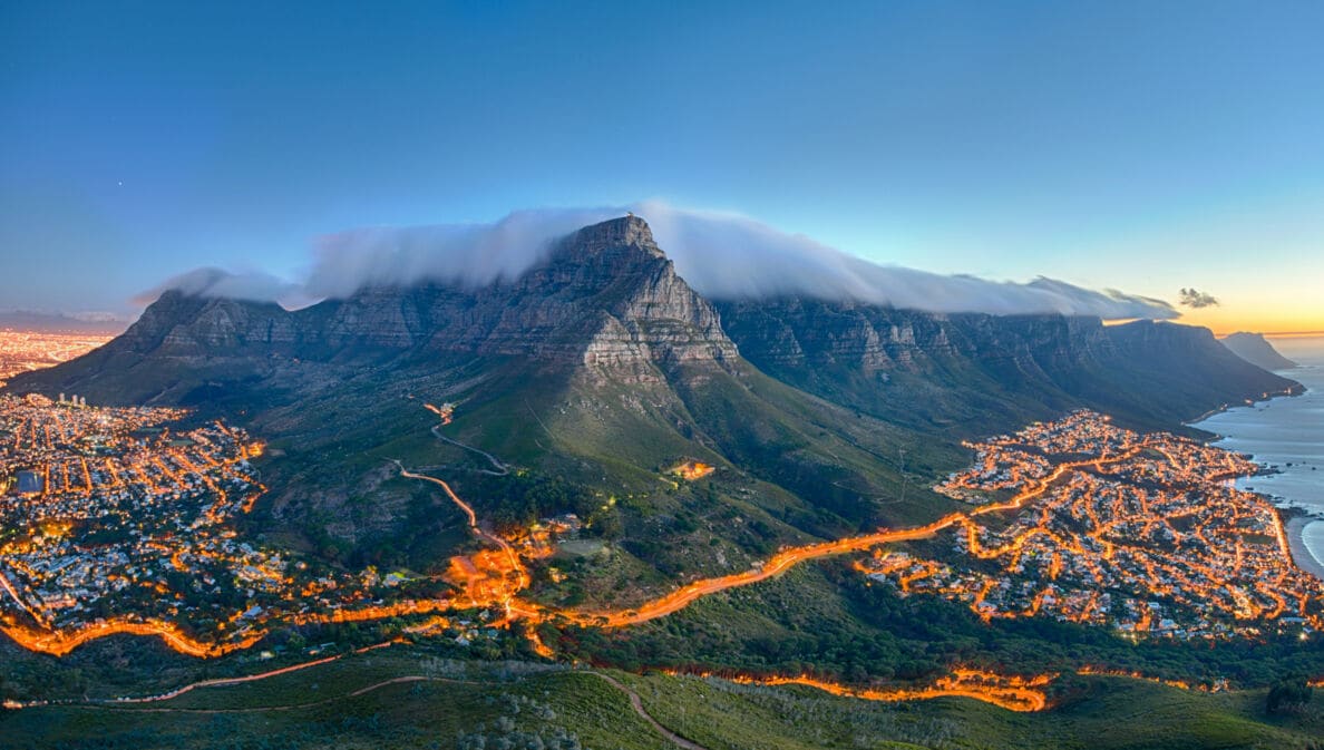 Tafelberg mit Wolkenkappe über Kapstadt bei Sonnenuntergang, beleuchtete Stadt und Küste im Hintergrund
