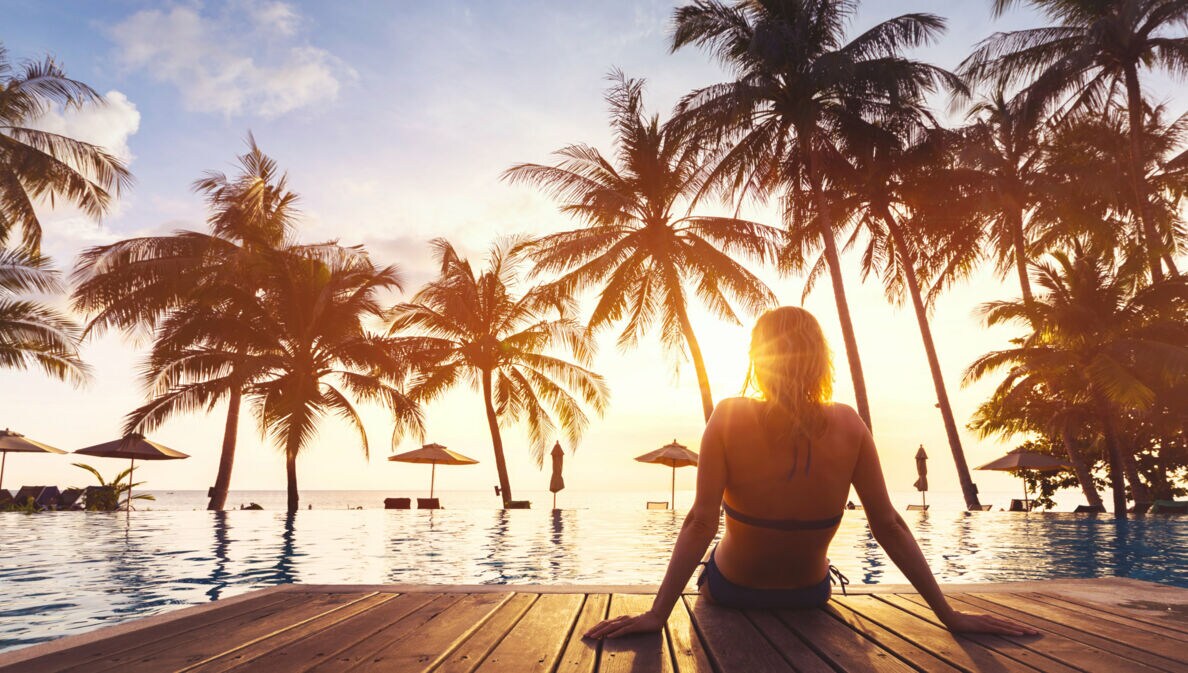 Frau sitzt am Rand eines Pools mit Blick auf Sonnenuntergang und Palmen