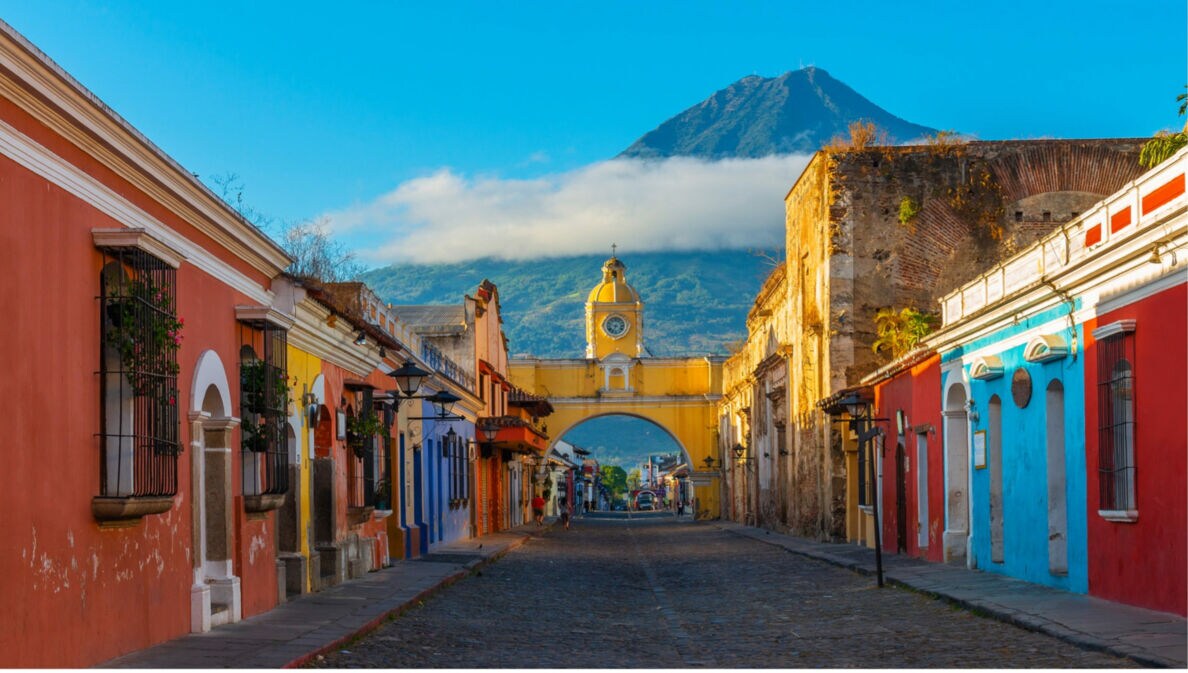 Blick auf die gelbe Santa-Catalina-Arkade in Antigua, Guatemala, mit Kopfsteinpflasterstraße und Vulkan Agua im Hintergrund