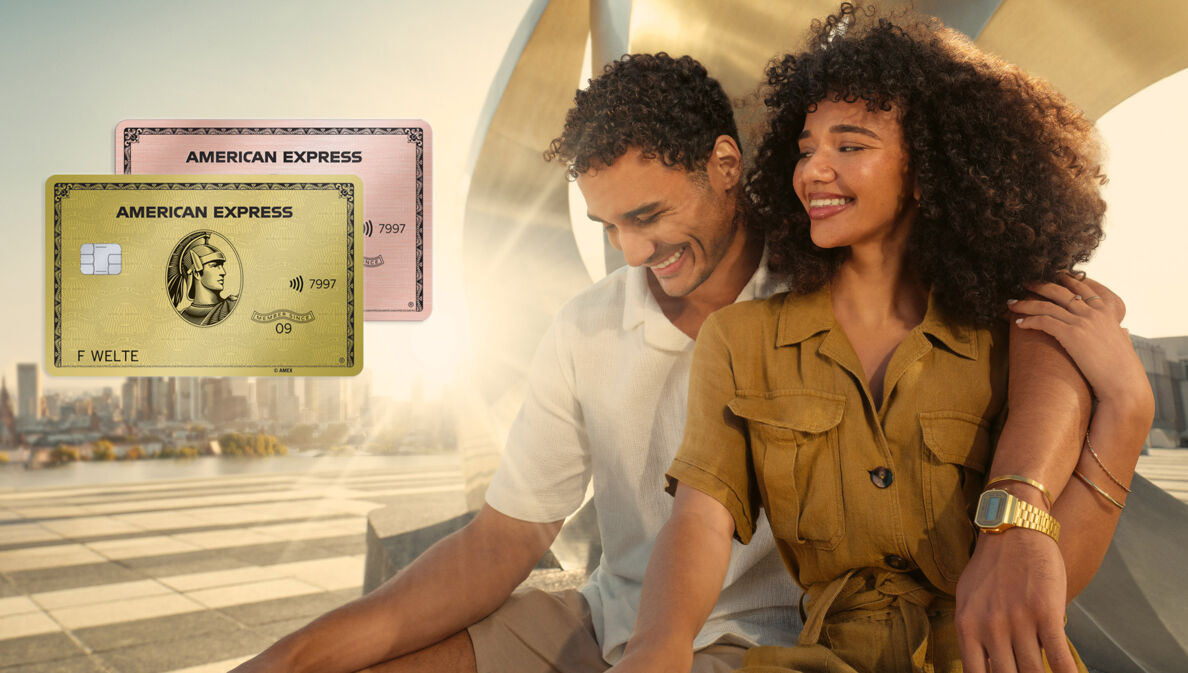 Ein lächelndes Paar sitzt vor einer gold glänzenden Metallskulptur im Sonnenschein auf einem weitläufigen Platz, er hat einen Arm um sie gelegt.