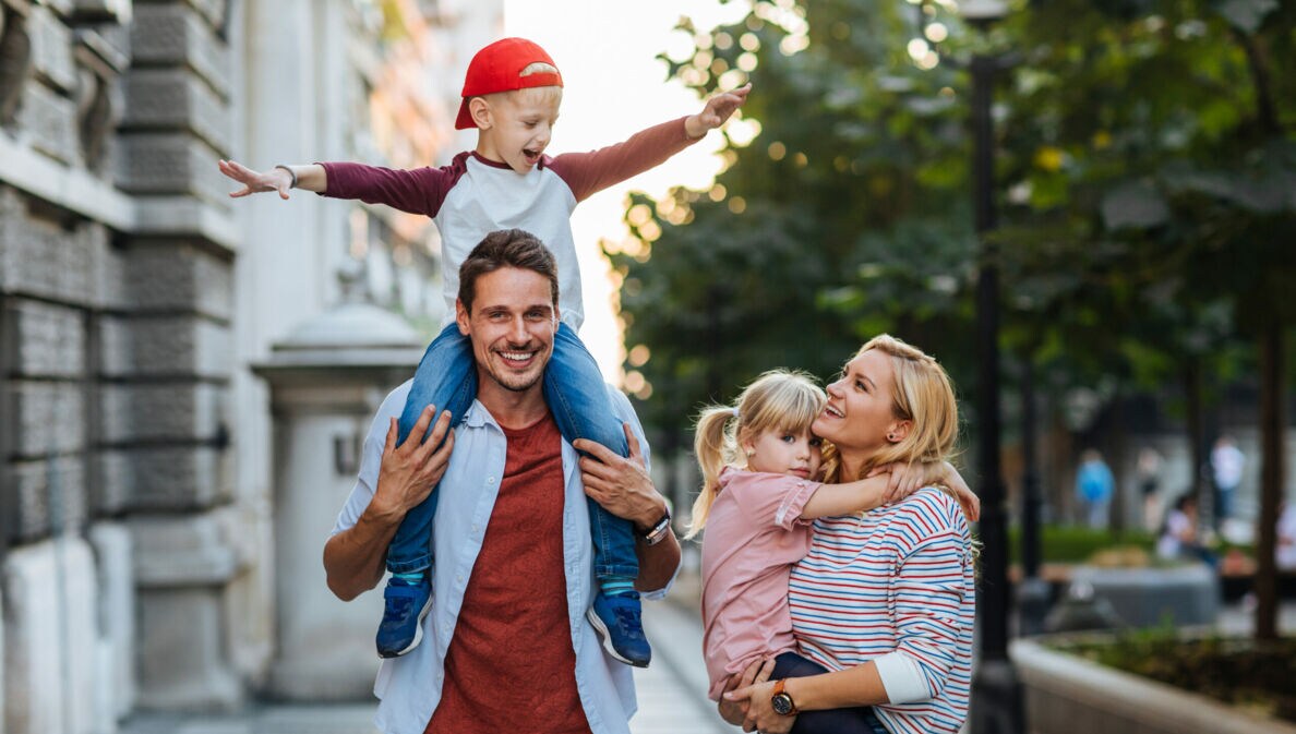 Ein Mann auf einem Weg zwischen Haus und Straße trägt einen Jungen auf den Schultern, eine Frau daneben trägt ein Mädchen.