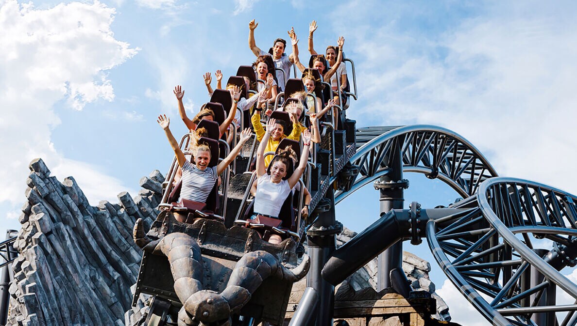 Menschen fahren auf der Achterbahn Taron im Phantasialand, Arme in die Luft gestreckt, vor blauem Himmel