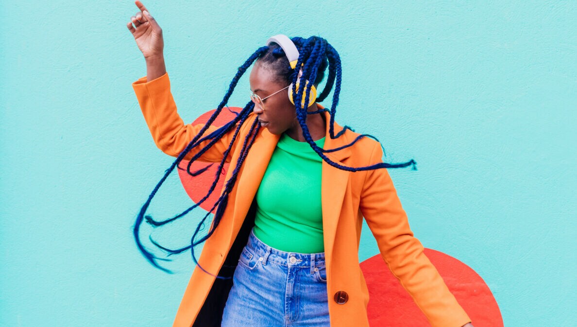 Eine junge Frau mit blauen Rastazöpfen in einem grünen Shirt und orangefarbenen Mantel tanzt mit gelben Kopfhörern vor einer türkisfarbenen Fassade.