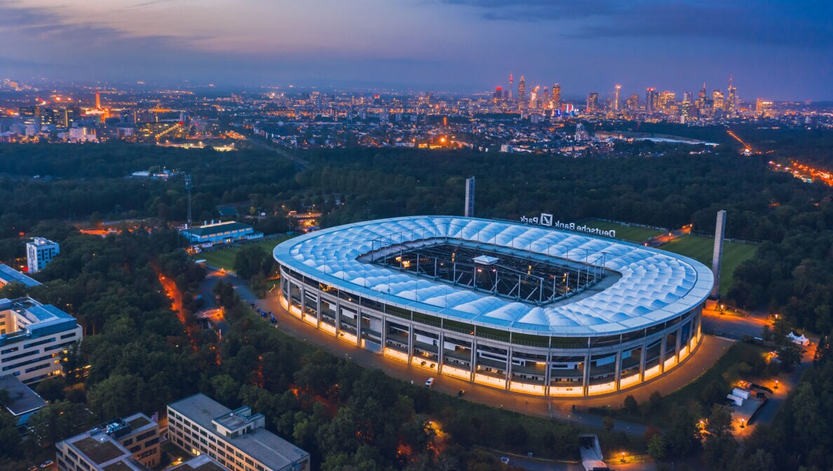 Luftaufnahme eines Fußballstadions, umgeben von Waldgebiet, vor der erleuchteten Skyline Frankfurts bei Nacht.