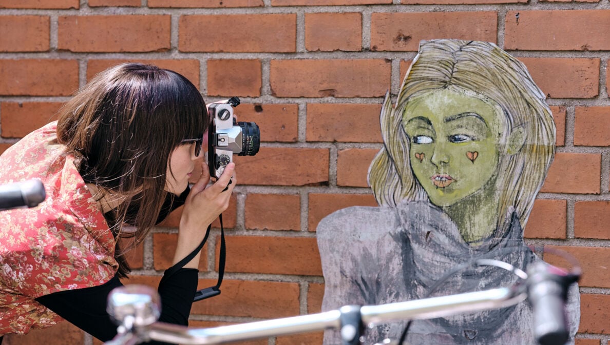 Frau mit braunen Haaren und rotem Blumenmuster-Oberteil fotografiert eine Wandzeichnung einer Person mit langen Haaren vor einer Backsteinmauer.