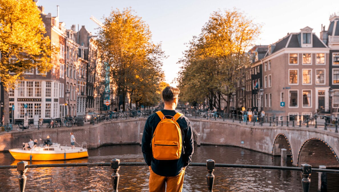 Rückansicht einer Person mit Rucksack, die an einem sonnigen Tag an einem Kanal in Amsterdam steht, auf der anderen Seite Häuser.