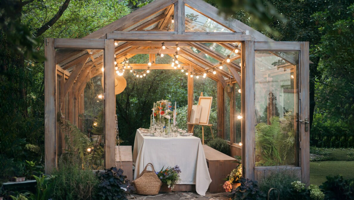 Holz-Gewächshaus mit Glaselementen, innen gedeckter Tisch mit Blumen und Kerzen, Lichterkette am Dach, umgeben von Gartenpflanzen