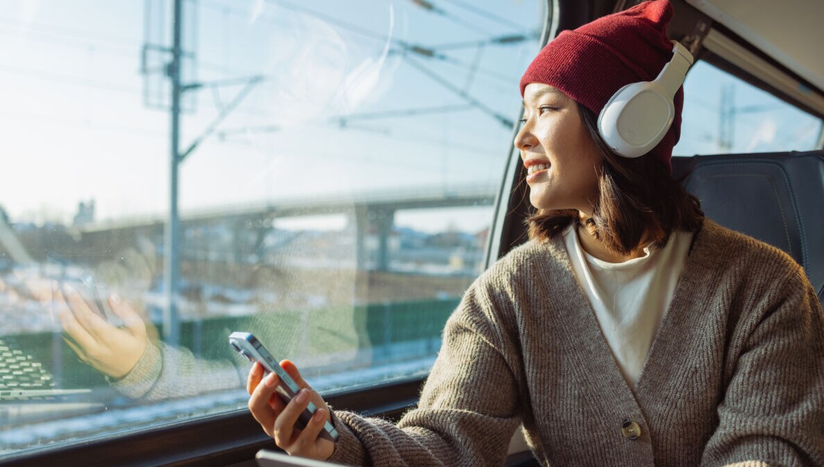 Eine Person mit Kopfhörer und Smartphone sitz in einem Zug. Sie trägt eine rote Mütze und einen grauen Pullover. Im Hintergrund sind Züge und Schienen zu sehen.