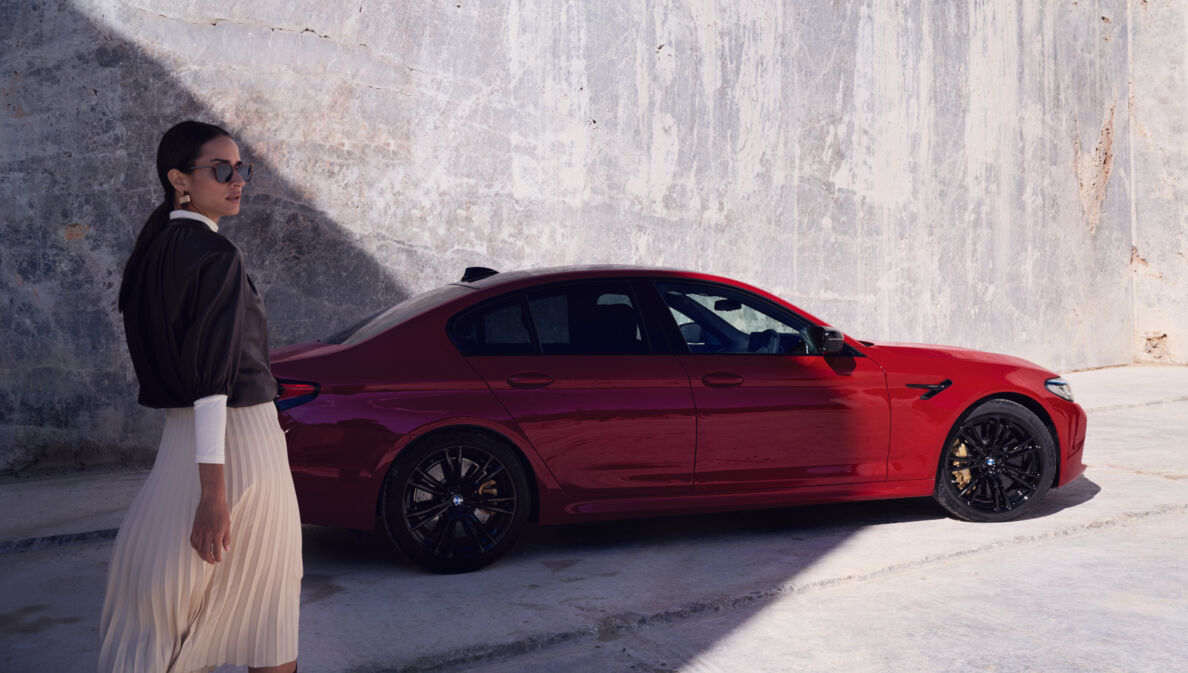 Eine elegante Frau mit Sonnenbrille vor einem roten BMW vor einer Betonwand.