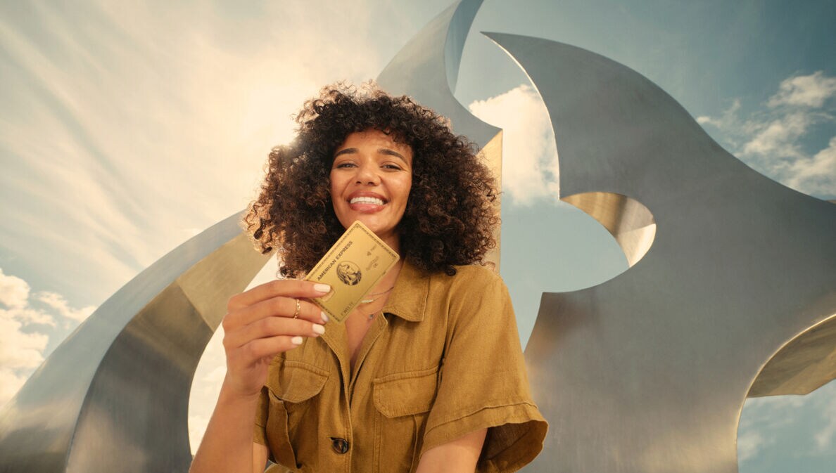 Eine lächelnde junge Frau hält eine goldene Kreditkarte von American Express in der Hand, im Hintergrund eine moderne Skulptur im Freien bei Sonnenschein.