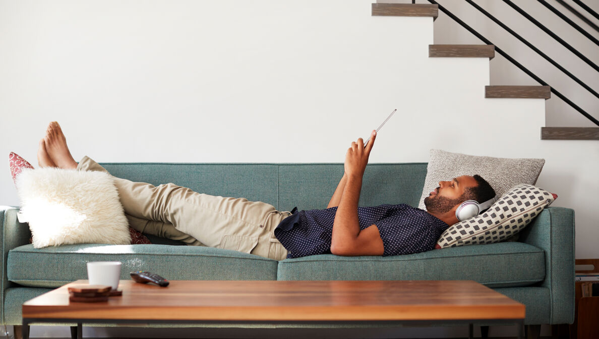 Ein Mann liegt mit Kopfhörern über den Ohren auf einem Sofa und liest.