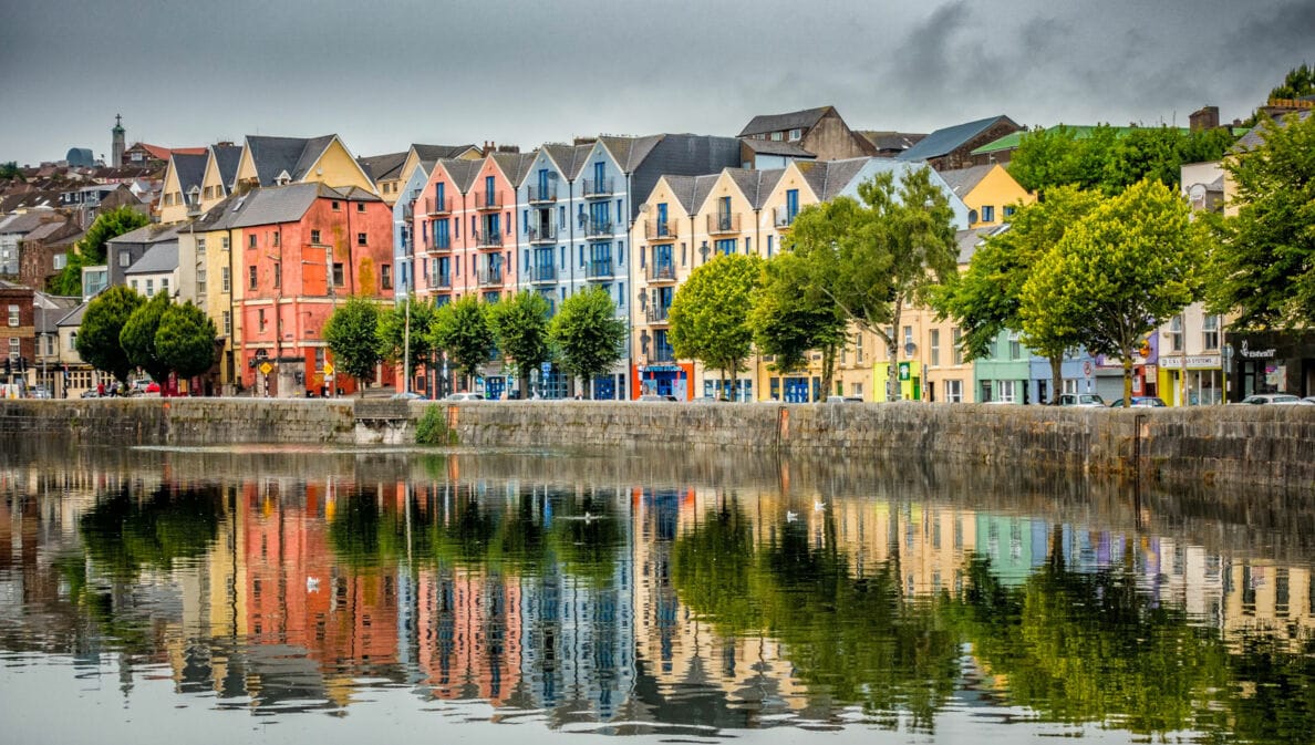 Bunte Wohngebäude in verschiedenen Farben spiegeln sich im ruhigen Wasser eines Hafens. Bäume stehen entlang der Uferpromenade.