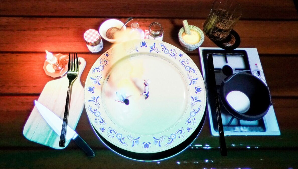 Ein gedeckter Tisch mit einem großen weißen Teller, auf dem zwei kleine Figuren stehen. Daneben befinden sich ein Messer, eine Gabel, ein Glas, Gewürzgläser und eine Pfanne mit einem Ei.