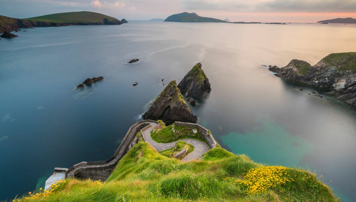 Blick auf eine Küstenlandschaft mit grünem Hügel, steilen Klippen und ruhigem Wasser. Im Hintergrund sind mehrere Inseln und ein Boot sichtbar.