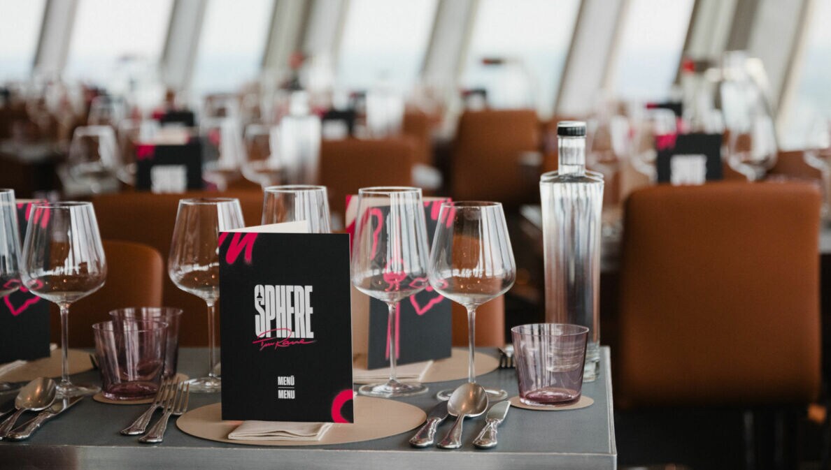 Tischgedeck in einem Restaurant mit Gläsern, einer Flasche und einer Speisekarte mit dem Titel 'SPHERE'. Im Hintergrund sind weitere Tische sichtbar.