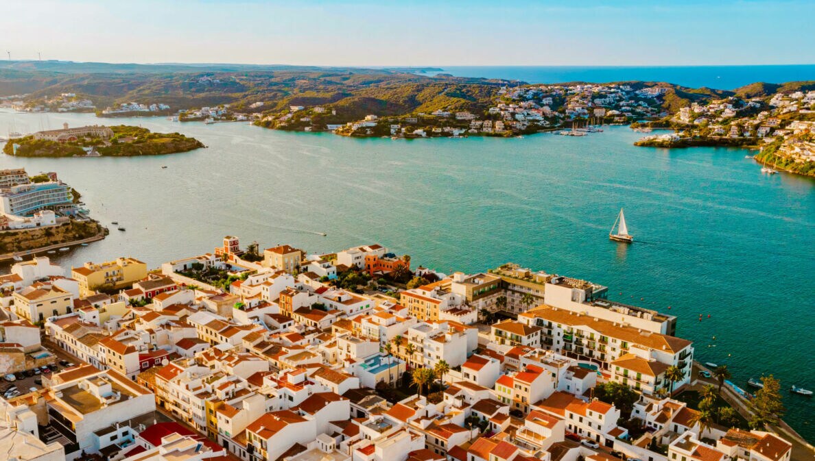 Luftaufnahme einer Küstenstadt mit weißen Gebäuden und roten Dächern, umgeben von Wasser und Hügeln. Ein Segelboot ist auf dem Wasser sichtbar.