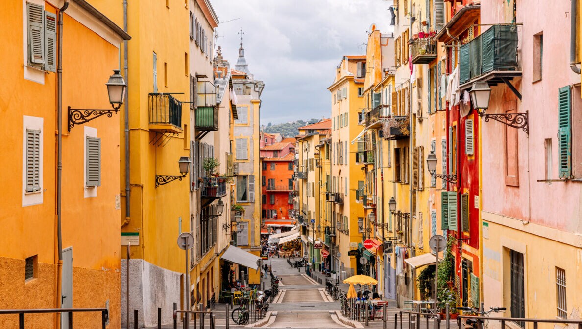 Blick auf eine enge, gepflasterte Straße mit bunten Gebäuden in Gelb- und Orangetönen. Auf beiden Seiten stehen Balkone mit Pflanzen. Im Hintergrund sind Tische und Stühle eines Cafés zu sehen.