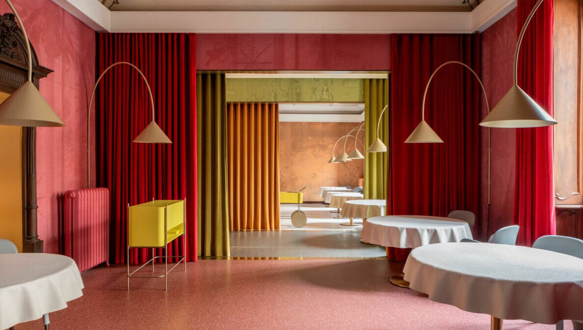 Ein eleganter Innenraum mit runden Tischen, die mit weißen Tischdecken bedeckt sind. An den Wänden hängen rote und grüne Vorhänge. Im Hintergrund sind gelbe und orangefarbene Wände sichtbar.