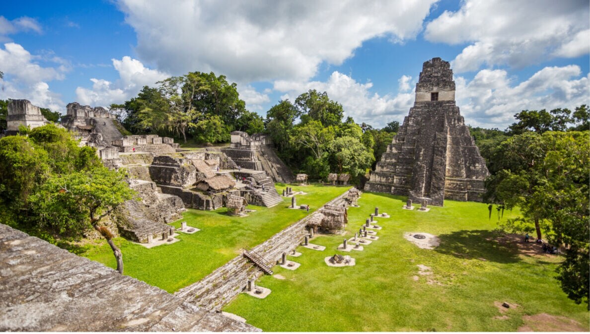 Maya-Tempelanlage mit Stufenpyramide und mehreren Steinstrukturen umgeben von grünem Gras und Bäumen unter blauem Himmel mit Wolken