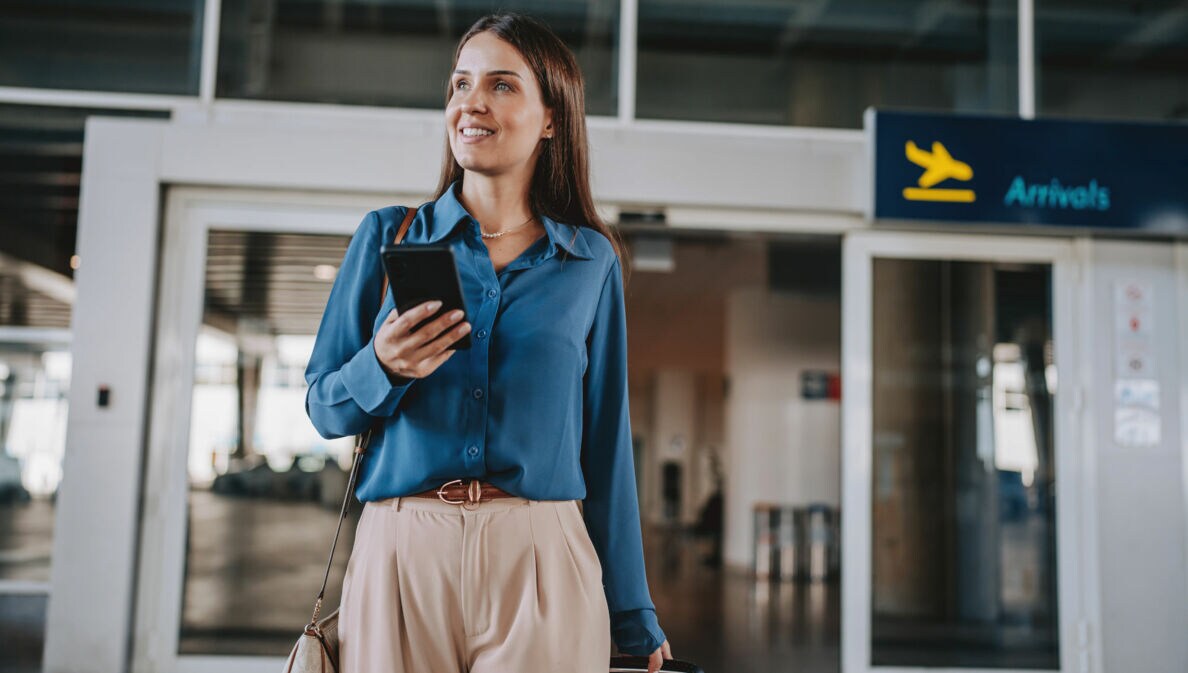 Eine elegant gekleidete Frau mit Smartphone steht vor dem Eingang der Ankunftshalle eines Flughafen-Terminals.