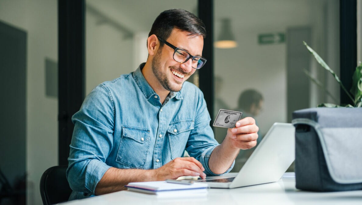 Ein gut gelaunter Mann im Jeanshemd sitzt an einem Tisch vor einem Laptop in einem Büro und hält die Business Platinum Card von American Express in der Hand.