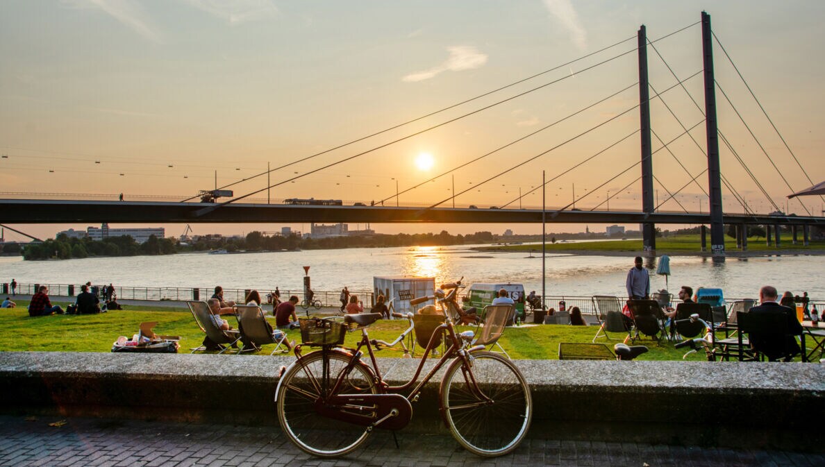 Ein rotes Fahrrad steht auf dem Bürgersteig, im Hintergrund ist eine Brücke und der Sonnenuntergang über dem Wasser zu sehen. Menschen entspannen auf der Wiese.