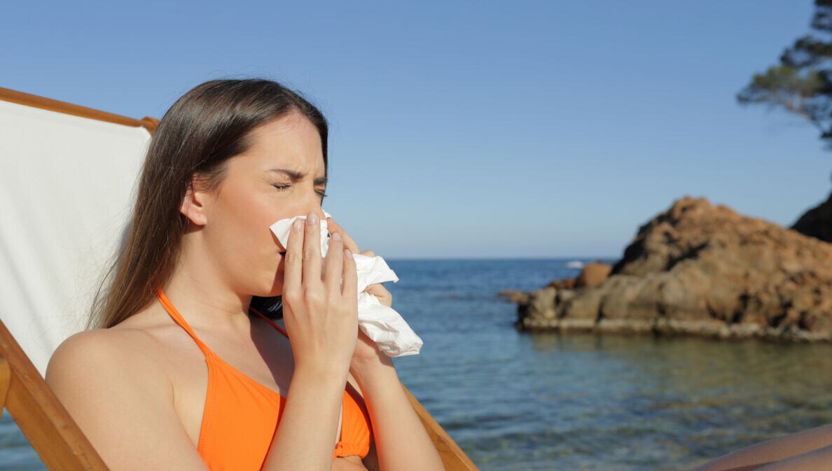 Eine Frau in einem orangefarbenen Bikini sitzt auf einer Liege am Strand und hält ein Papiertaschentuch in der Hand. Im Hintergrund sind Felsen und das Meer sichtbar.