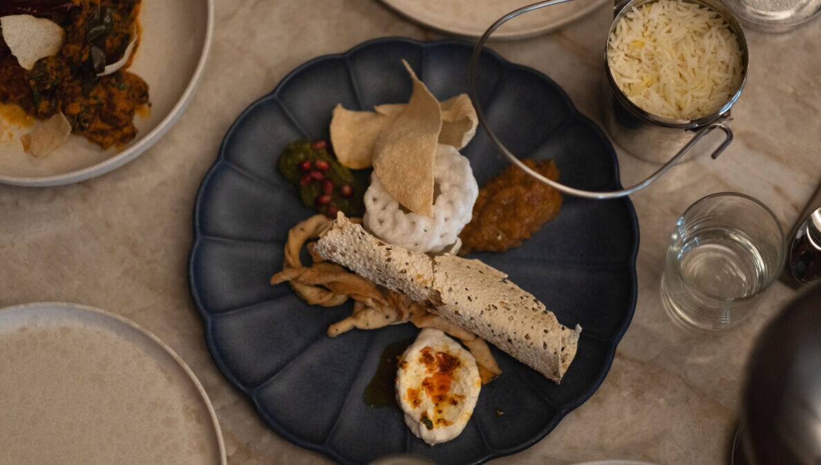 Ein blauer Teller mit verschiedenen indischen Gerichten: Reis in einer Schüssel, Papadam, Chutney, frittiertes Gemüse und ein gerolltes Fladenbrot.