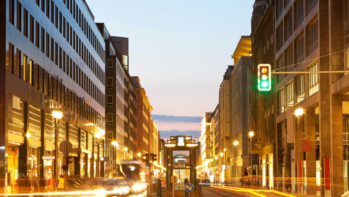 Fotografie mit langer Belichtungszeit einer befahrenen Straße während der Abenddämmerung, der Fokus liegt auf einer grünen Ampel.