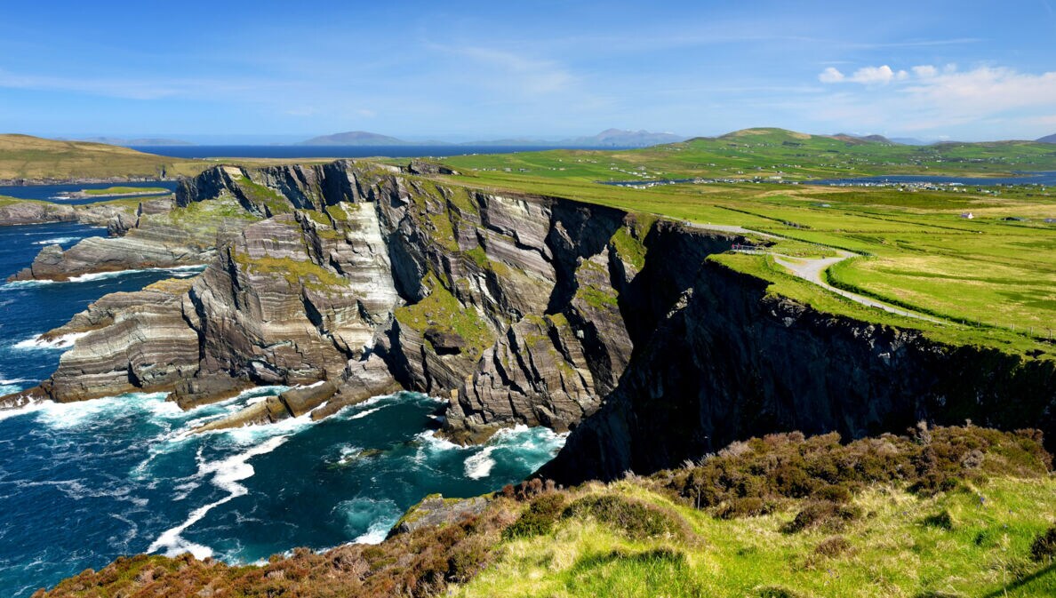 Luftaufnahme einer Küstenlandschaft mit steilen Klippen, grünem Gras und einem schmalen Weg, der entlang der Klippen verläuft. Im Hintergrund sind sanfte Hügel und ein blauer Himmel zu sehen.