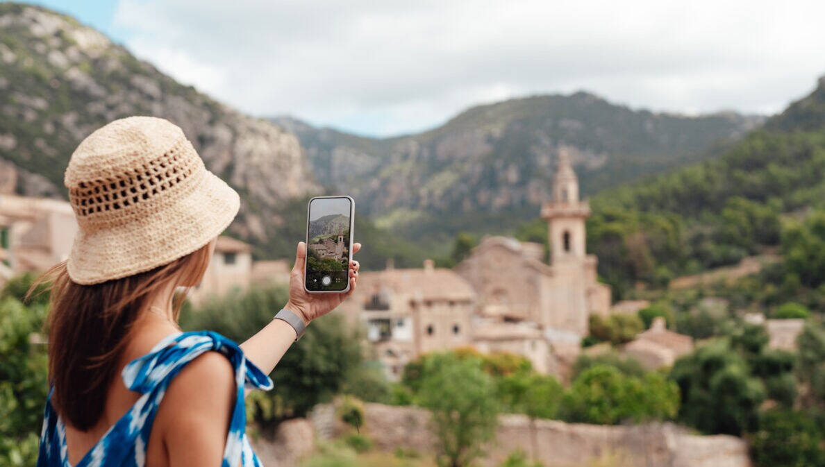 Frau mit Strohhut und blauem Kleid fotografiert Bergdorf mit Smartphone