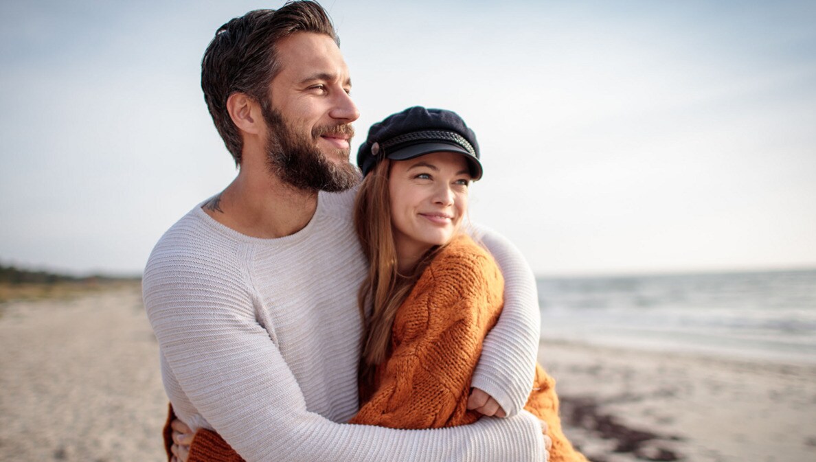 Ein sich umarmendes Paar in Pullovern steht glücklich lächelnd an einem Sandstrand und blickt aufs Meer.