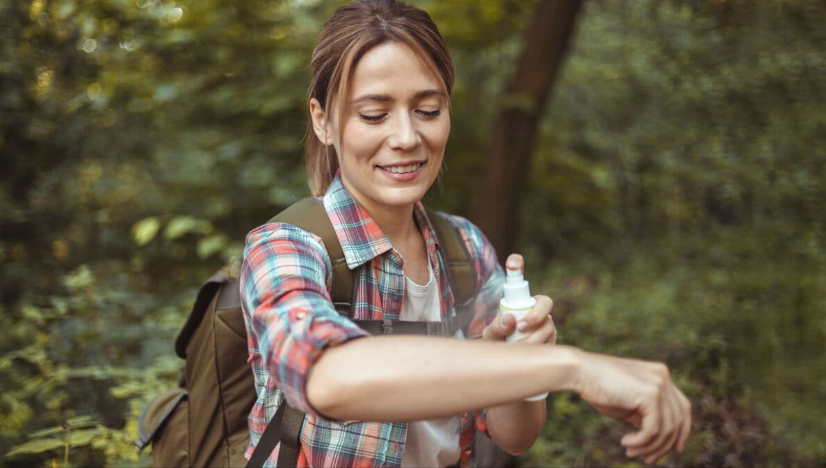 Person mit kariertem Hemd und Rucksack trägt im Wald Insektenschutzspray auf den Arm auf.