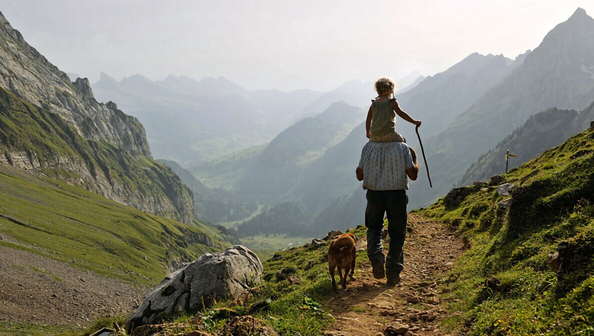 Ein Erwachsener trägt ein Kind auf den Schultern, während sie einen Wanderweg entlanggehen. Ein Hund läuft neben ihnen. Die Landschaft zeigt Berge und Täler im Hintergrund.