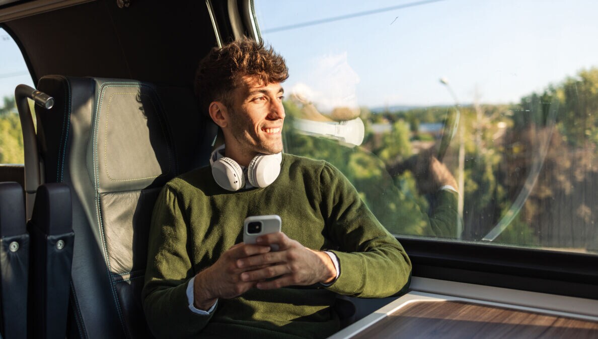 Junger Mann mit grünem Pullover und weißen Kopfhörern sitzt im Zug mit Smartphone in der Hand und blickt aus dem Fenster.
