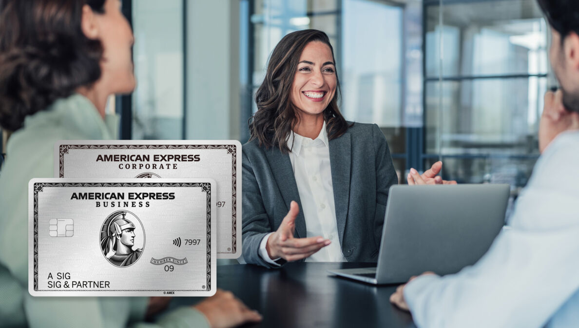 Eine lachende Frau im grauen Blazer sitzt vor einem Laptop an einem Tisch gegenüber zwei Personen in einem Raum mit Glaswänden.