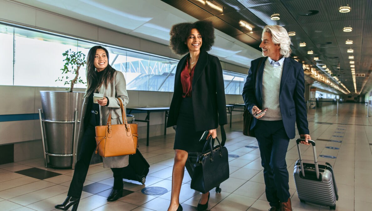 Drei Personen in eleganter Business-Kleidung mit Handgepäck laufen nebeneinander durch ein Flughafenterminal.