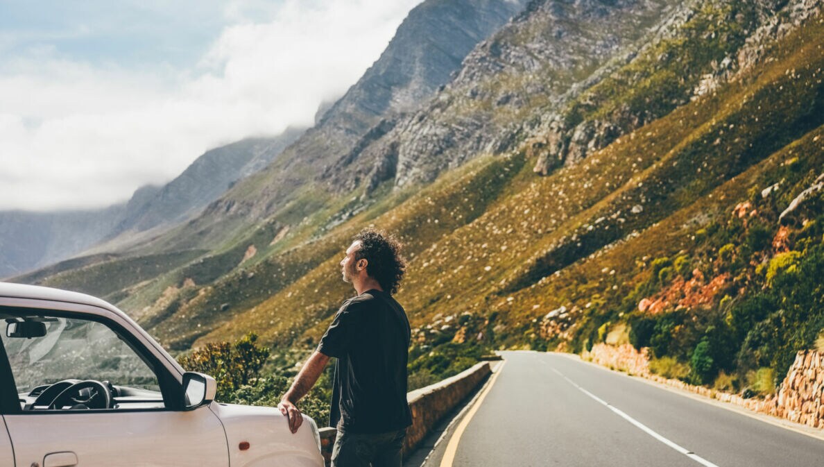 Mann lehnt an weißem Auto an einer Bergstraße, umgeben von steilen Felsen und bewölktem Himmel.