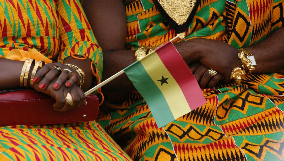 Oberkörper zweier sitzender Frauen in traditionellen ghanaischen Kleidern aus buntem Kente-Stoff und Goldschmuck, davor eine kleine ghanaische Flagge aus Papier in einer Hand gehalten.