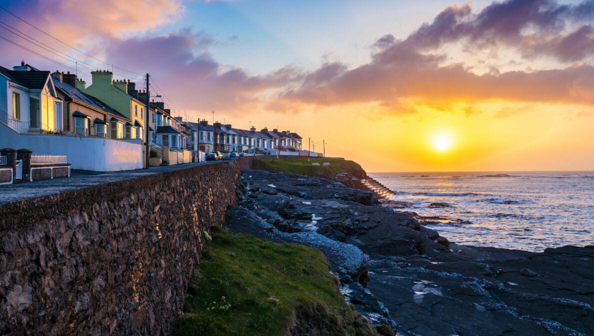 Sonnenuntergang über der Küste von Kilkee mit Reihenhäusern auf einer Steinmauer und Felsen am Meer
