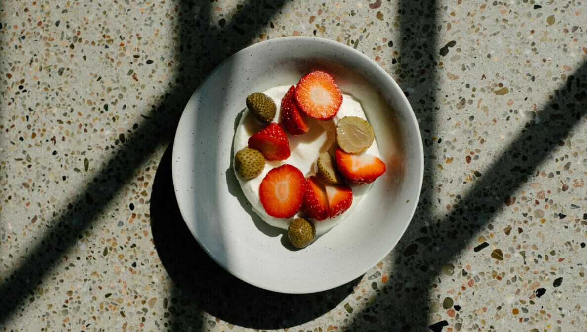 Schale mit Joghurt, belegt mit geschnittenen Erdbeeren und kleinen grünen Früchten, auf marmoriertem Untergrund mit Schatten.