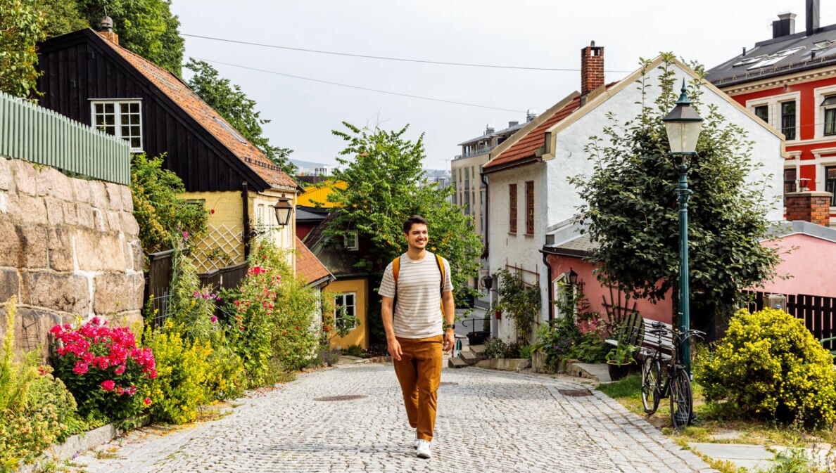 Person mit gestreiftem T-Shirt und brauner Hose geht auf gepflasterter Straße in Oslo, umgeben von bunten Häusern und Blumen.