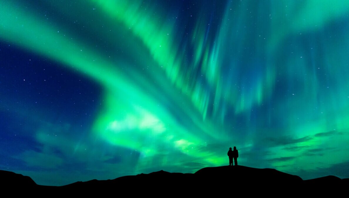 Zwei Personen stehen auf einem Hügel und beobachten das grün leuchtende Nordlicht am Nachthimmel in Norwegen.