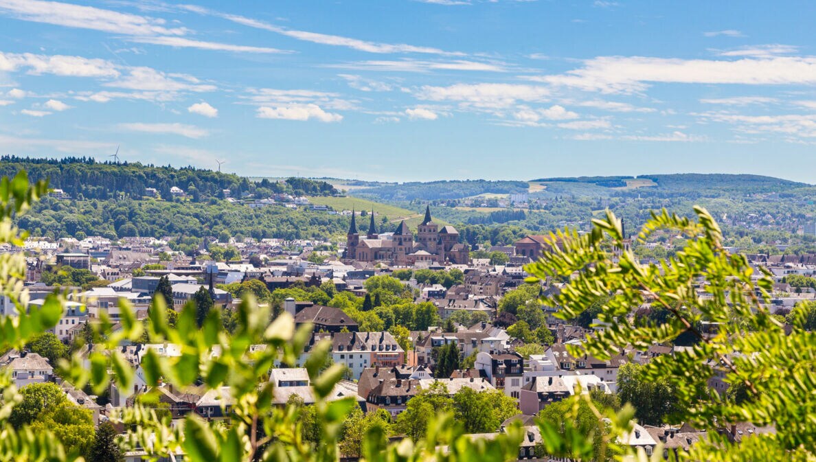 Blick auf Trier mit der Porta Nigra und umliegenden Gebäuden, im Vordergrund grüne Blätter, im Hintergrund bewaldete Hügel unter blauem Himmel mit Wolken.