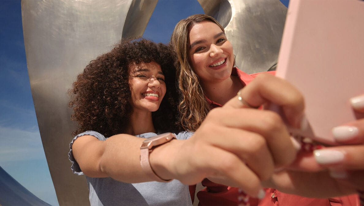 Zwei junge Frauen machen ein Selfie mir einem rosafarbenen Smartphone vor einer Metallskulptur unter freiem Himmel.