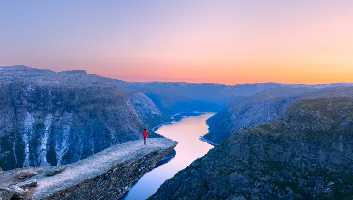 Bergklippe Trolltunga mit Person in roter Jacke, die auf dem Felsvorsprung über einem tiefen Fjord steht, umgeben von Bergen im Abendlicht.