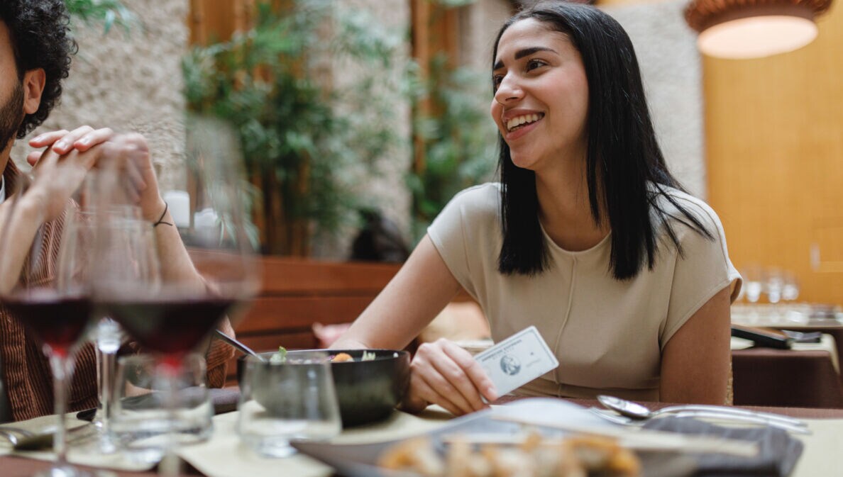 Eine Frau sitzt neben einer anderen Person an einem gedeckten Tisch und hält eine Platinum Plus Card von American Express in der Hand.
