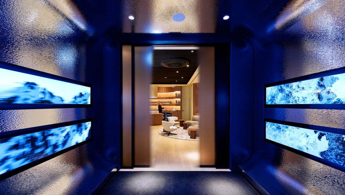 Blick durch einen dunklen, blau beleuchteten Gang mit vier rechteckigen Bildschirmen an den Seiten auf einen hellen, modernen Raum mit Sesseln und Regalen.