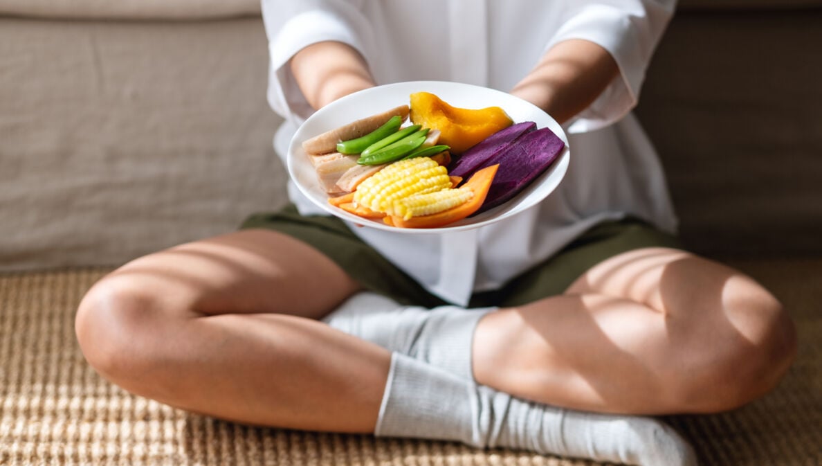 Person sitzt im Schneidersitz und hält einen Teller mit buntem, geschnittenem Gemüse, darunter Mais, Süßkartoffel und grüne Bohnen.